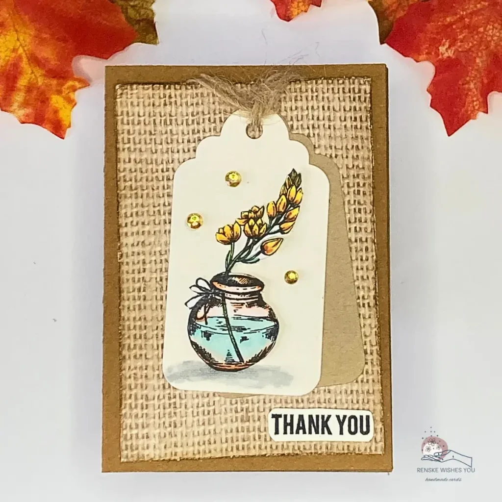 A top-down view of an autumn thank you card featuring a kraft card base with a burlap-style background, layered white and brown tags, a softly coloured floral vase as the focal image, a few yellow rhinestones for detail, and a small “Thank You” sentiment tucked into the lower right corner.