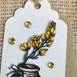 A close-up of three small yellow rhinestones placed around the floral image on the white tag, with two on the left side and one on the right to add a balanced autumn accent.