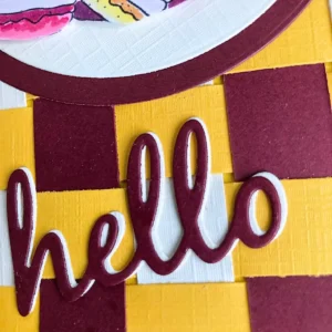 Close-up of the die-cut “Hello” sentiment on the handmade fall card, shown at a slight angle to highlight the layered shadow effect. The top word is wine red, offset over a light yellow layer that creates a subtle shadow beneath the letters. The photo captures the clean edges of the die-cuts and the soft fall tones that match the rest of the card.
