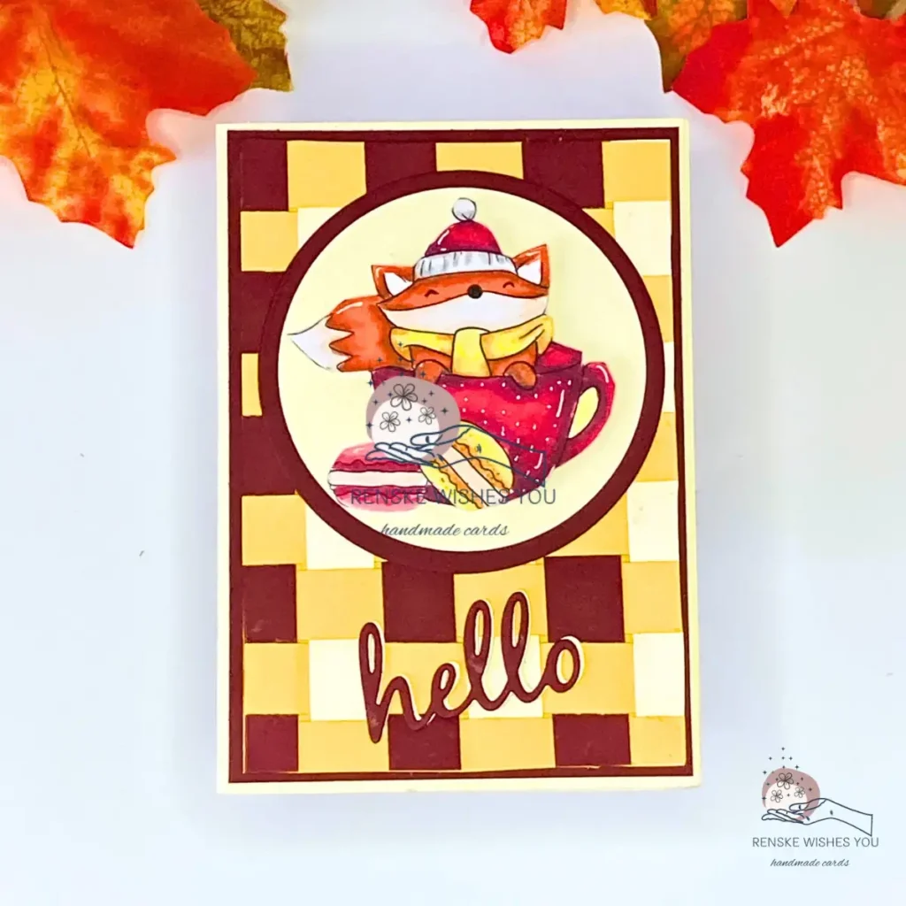Handmade fall card featuring a fox in an autumn cup, layered on a woven paper background in yellow, cream, and wine-red tones. The card is photographed straight on, with autumn leaves visible in the top corners. The design shows warm colours, circle layers, and a ‘Hello’ sentiment