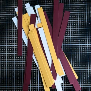 Stack of paper strips prepared for the woven background of a handmade fall card. The strips are cut in yellow, cream, and wine-red colours. The photo shows the paper neatly arranged and ready for weaving