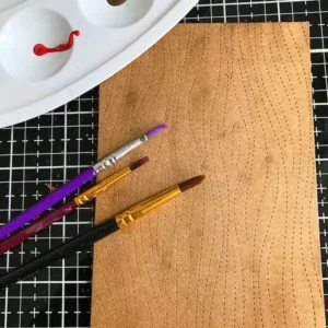 Ink-blended woodgrain background with a paint palette and three brushes in the background, showing the creative process of the autumn card tutorial.