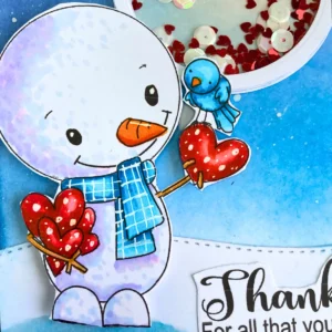 Close up colored snowman with ohuhu markers Close-up of the coloured digital stamp, showing detailed alcohol marker shading: snowman in soft blues, pink scarf, celadon blue accents, orange nose, brown arms, and small red hearts on the chest. Gel pen highlights add dimension to the scarf and hearts.