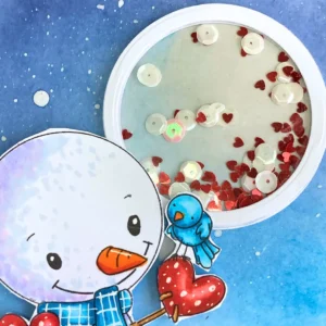 Close up of the moon shaker Close-up of the moon shaker element for a winter thank-you card, showing white layered circles, a transparent acetate window, and red heart and white circle shaker pieces inside. Distress Ink on the back circle creates a glowing moon effect.