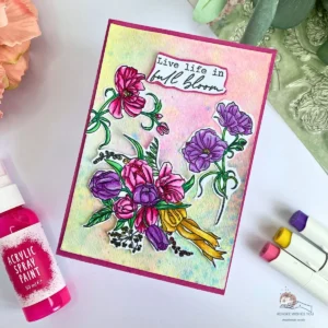 Handmade spring card with flowers photographed at an angle, showing a soft watercolour background in blue, pink and yellow with layered floral elements and sentiment