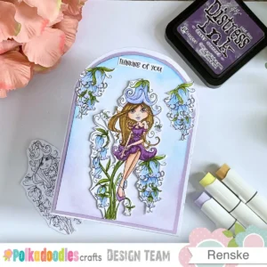 A completed handmade thinking of you card sits on a white surface next to soft peach flowers and markers. The card features a rounded arch shape at the top with a hand coloured girl digital stamp in the centre. She is surrounded by clusters of blue flowers against a soft blue and purple background.