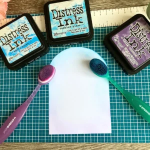 Three square distress ink pads in blue and purple shades are arranged around a white arched card panel. Two blending brushes with colourful bristles lie ready on a green cutting mat for the background. This setup shows how to start the ink blending for an arch card tutorial.