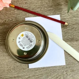 A small metal bowl sits upside down on a white cardstock square to help trace a curve. A red pencil and a white bone folder lie on the wooden table next to the paper. This achievable method shows how to make an arch shaped card without using special dies.
