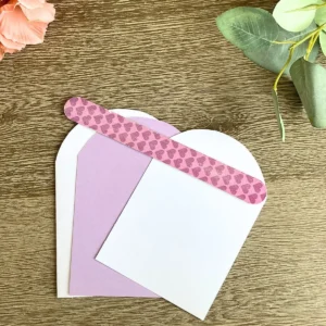 Three arched paper panels in white and lilac are stacked slightly apart on a wooden table. A pink nail file with a heart pattern lies across the top of the cardstock layers. This tool is used to sand the edges for a smooth and peaceful finish on your project.
