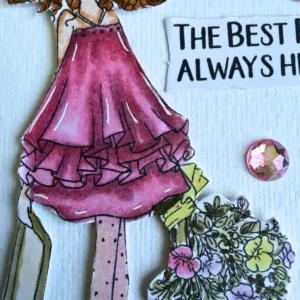 A close-up photograph showing the ruffled pink dress of the coloured Ruby digital stamp. The dress features multiple frills shaded in rose and magenta alcohol markers with fine white highlight lines . Placed on the white textured paper next to the dress is a round pink crystal gem . The top of a pastel bouquet and part of a paper sentiment banner rest beside the ruffle