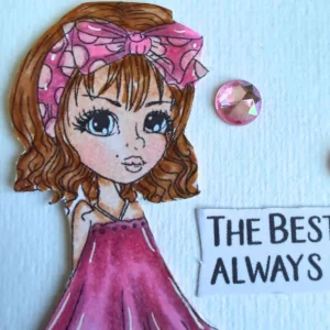 A close-up photograph focusing on the coloured face of the Ruby digital stamp character. She has wavy brown hair tied with a pink polka-dot bow, big blue eyes, and subtle cheek shading coloured with alcohol markers . Positioned on the white textured paper beside her face is a round pink crystal gem and part of a printed paper sentiment banner.
