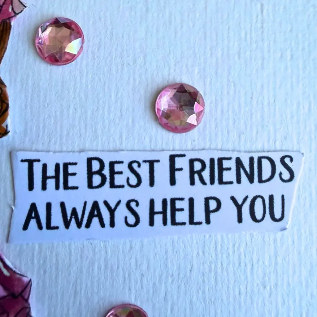 A close-up photograph of the printed sentiment banner reading the best friends always help you in clean black capital letters . The trimmed white paper strip sits on a textured cream cardstock panel . Two round faceted pink crystal gems are placed above and below the text