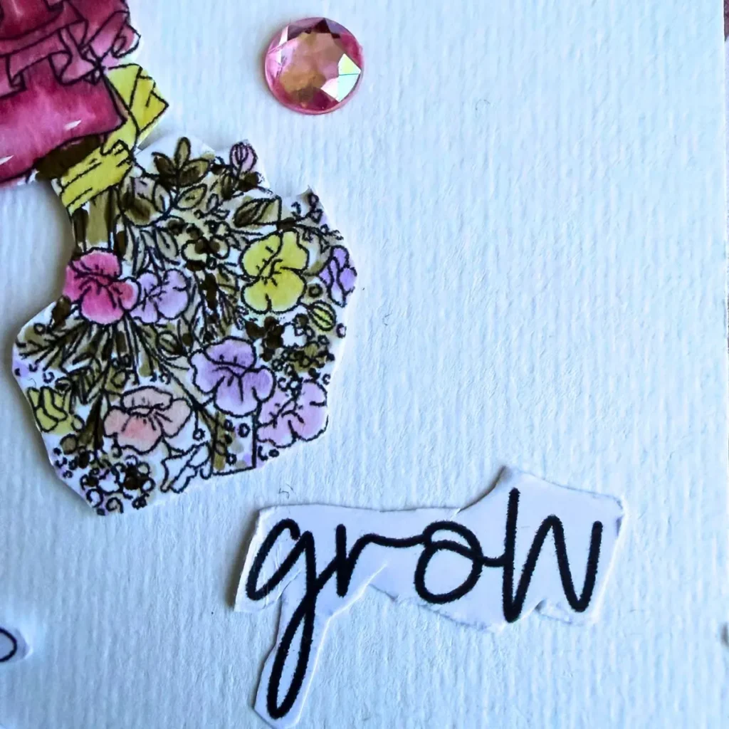 A close-up photograph of the sentiment word grow printed in black brush script on a white cut-out paper strip . The sentiment rests on a textured cream cardstock surface . It is positioned beneath a coloured pastel flower bouquet and a round pink crystal rhinestone