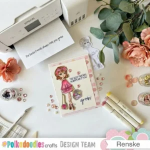 A top-down view of a crafting table with a completed handmade friendship card set in the middle . The card includes a coloured illustration of a girl in a pink tiered dress holding a bouquet . Round pink gems and printed sentiment banners reading the best friends always help you grow rest on the panel . Surrounding supplies on the desk include an Epson printer with printed text, Ohuhu markers, glass jars with pink embellishments, peach flowers, and a craft knife on a cutting mat.