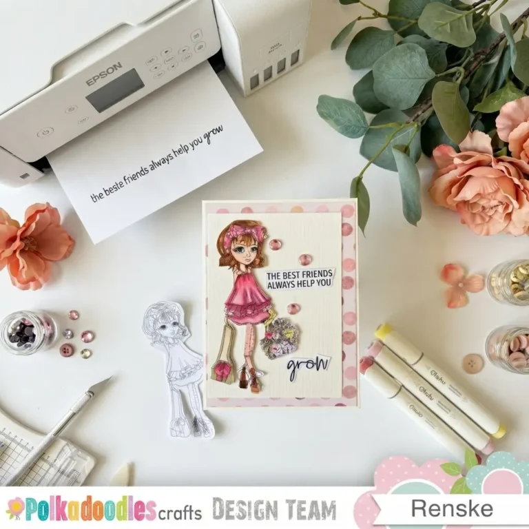 A flat lay top-down photograph of a craft workspace featuring a finished handmade friendship card in the centre . The card shows a coloured girl character in a frilly pink dress holding a floral bouquet . Round pink gems, a polka-dot border, and paper sentiment banners surround the main illustration . On the white table lie Ohuhu alcohol markers, glass jars with pink sequins, peach flowers, green leaves, and a craft blade . An Epson printer sits in the background with a printed sentiment strip.