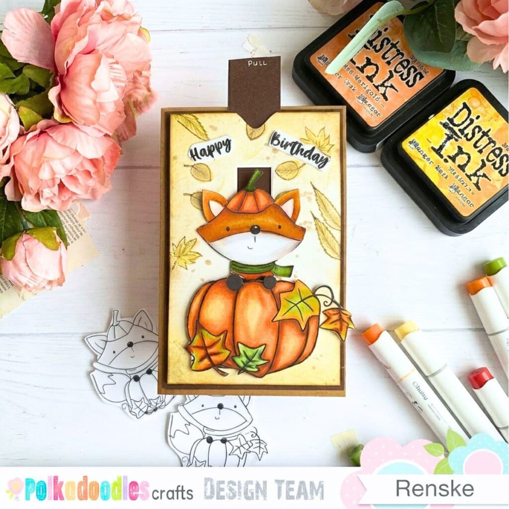 How to make a slider card with an autumn fox, featuring an orange fox peeking over a large orange pumpkin decorated with colourful autumn leaves. The cream background has yellow-brown inked edges, leaf details and scattered splatters. The “Happy Birthday” sentiment sits at the top, with a brown pull tab labelled “Pull” extending above the card.